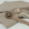 Woven pacifier clip with blush-and-ivory strap attached to two beige pacifiers